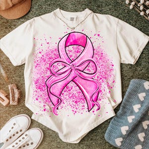 May include: Cream-colored t-shirt featuring a large pink ribbon design with a bow, surrounded by pink splatter. The ribbon has a leopard print pattern. The shirt is paired with white sneakers, a brown hat, and a blue blanket.