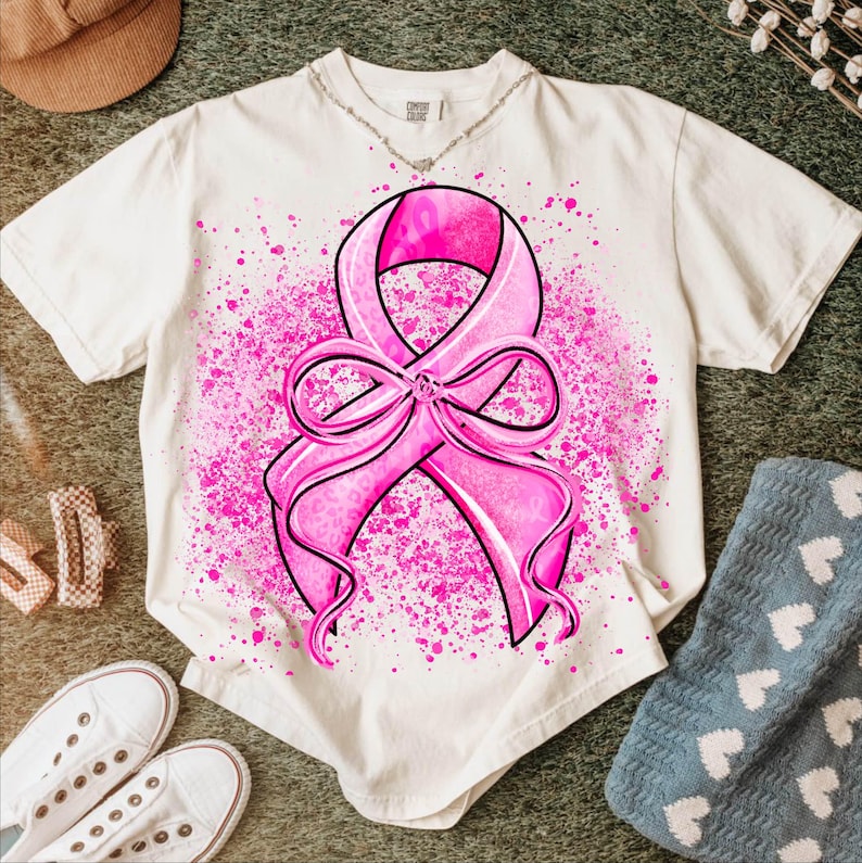 May include: Cream-colored t-shirt featuring a large pink ribbon design with a bow, surrounded by pink splatter. The ribbon has a leopard print pattern. The shirt is paired with white sneakers, a brown hat, and a blue blanket.