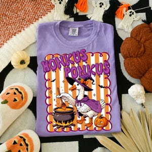 May include: A lavender t-shirt with a Halloween-themed graphic. The graphic features a goose in a witch hat and cape, stirring a cauldron, with the words "Honkus Ponkus". The design includes orange and white stripes, bats, and a jack-o'-lantern.