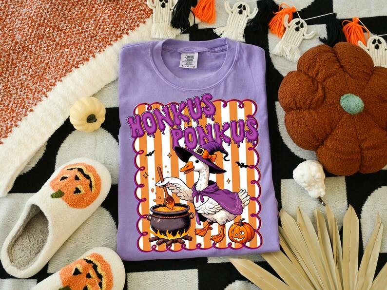May include: A lavender t-shirt with a Halloween-themed graphic. The graphic features a goose in a witch hat and cape, stirring a cauldron, with the words "Honkus Ponkus". The design includes orange and white stripes, bats, and a jack-o'-lantern.