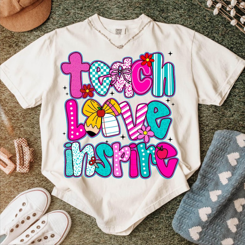May include: Cream-colored t-shirt with the words "Teach Love Inspire" in a colorful, patterned design. The design includes flowers, a pencil, and an apple. The shirt is surrounded by casual accessories.