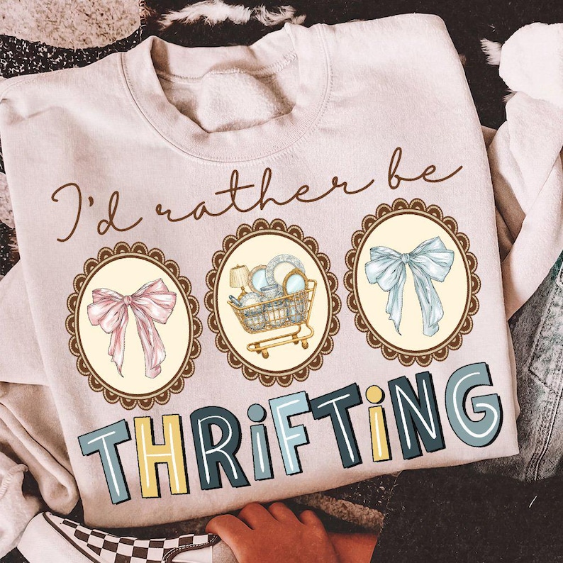 May include: A light beige sweatshirt with the phrase "I'd rather be thrifting" in brown script. The design includes three oval frames with a pink bow, a shopping cart filled with dishes, and a blue bow. The word "THRIFTING" is in large, colorful letters.