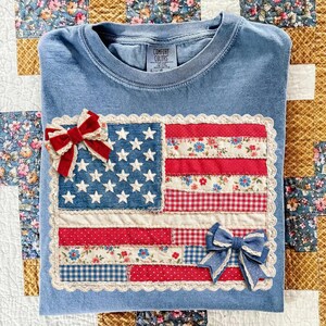 Puede incluir: Una camiseta azul Comfort Colors con un dise&ntilde;o de bandera estadounidense de patchwork. La bandera incluye rayas rojas y blancas, un campo azul con estrellas blancas y est&aacute; adornada con dos lazos decorativos.