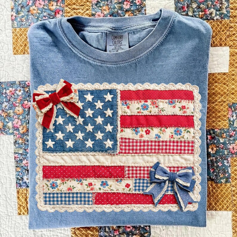 Puede incluir: Una camiseta azul Comfort Colors con un dise&ntilde;o de bandera estadounidense de patchwork. La bandera incluye rayas rojas y blancas, un campo azul con estrellas blancas y est&aacute; adornada con dos lazos decorativos.