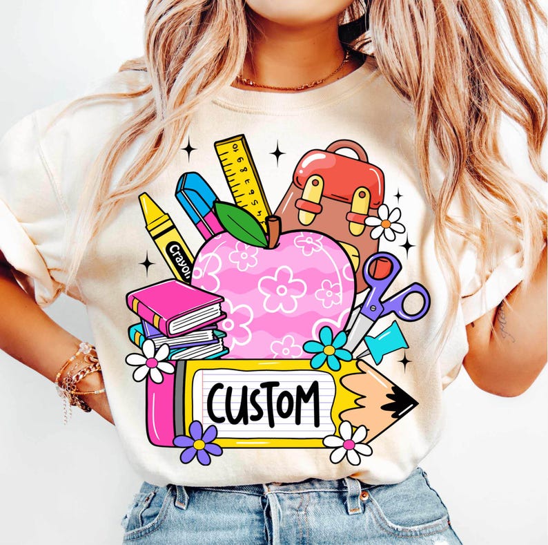 以下が含まれることがあります： クリーム色のTシャツ。カラフルな学校をテーマにしたグラフィックが特徴です。デザインには、花のアクセントが付いたピンク色のリンゴ、本、クレヨン、定規、そして「CUSTOM」という言葉が黄色い看板に描かれています。グラフィックは花と鉛筆で囲まれています。