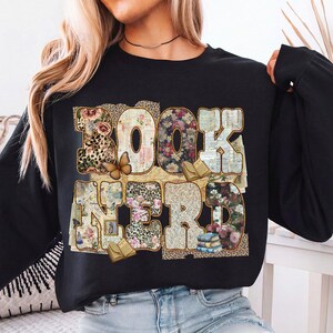 May include: Black sweatshirt with the words "BOOK NERD" in a collage of floral, leopard print, and book page patterns. A butterfly and small books are also featured in the design. The sweatshirt is a comfortable, casual style.