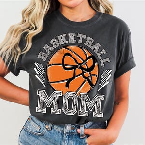 Pode incluir: Camiseta cinza escuro com um gr&aacute;fico de basquete. O design apresenta uma bola de basquete laranja com um la&ccedil;o preto, as palavras "BASKETBALL MOM" em negrito e detalhes de raios.