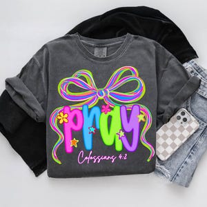 Puede incluir: Camiseta gris oscuro con la palabra "PRAY" en letras coloridas, adornada con un lazo arco&iacute;ris y detalles florales. El texto "Colosenses 4:2" est&aacute; debajo. La camiseta se muestra con ropa negra doblada y pantalones cortos de mezclilla.