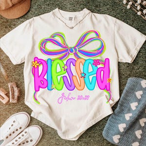 Puede incluir: Camiseta de color crema con un dise&ntilde;o gr&aacute;fico colorido. El dise&ntilde;o presenta la palabra "BLESSED" en colores vibrantes, un lazo decorativo y el texto "John 20:29". Es una prenda de vestir informal.