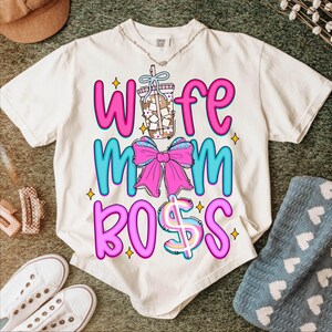 May include: Cream-colored t-shirt with the words "Wife Mom Boss" in pink and blue, featuring a bow and a drink graphic. The shirt has a casual, relaxed fit and is likely made of soft material.
