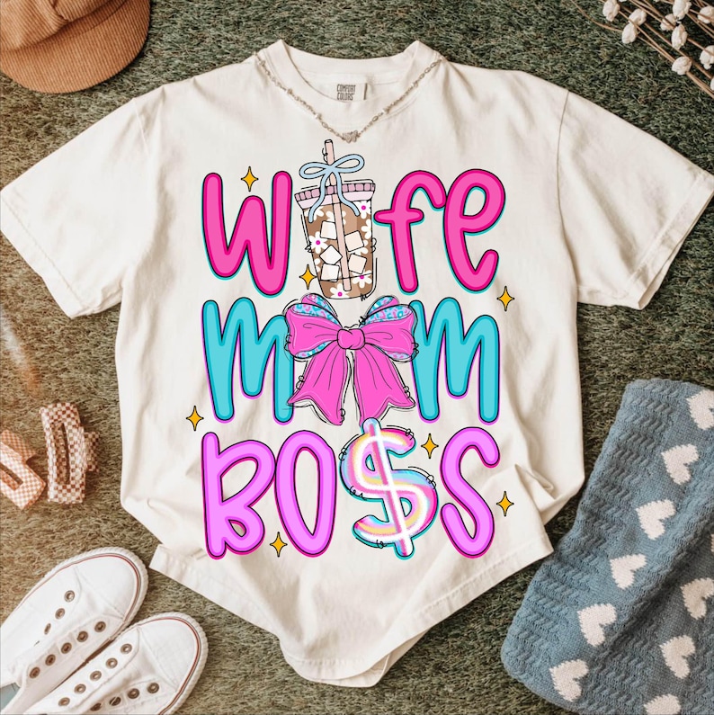 May include: Cream-colored t-shirt with the words "Wife Mom Boss" in pink and blue, featuring a bow and a drink graphic. The shirt has a casual, relaxed fit and is likely made of soft material.
