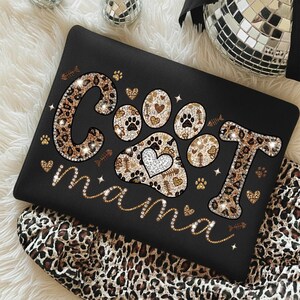 May include: Black tote bag with the words "Cool Mama" in a leopard print design, with paw prints and hearts. The bag is decorated with glitter and small fish bone and paw print accents.