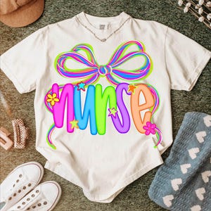May include: Cream-coloured t-shirt featuring a colourful bow design above the word "NURSE". The word is in bright, multi-coloured letters, with star and flower accents. A casual tee, perfect for nurses and healthcare workers.