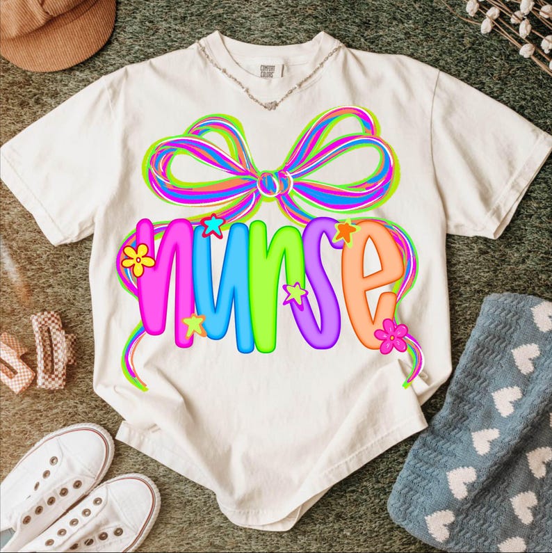 May include: Cream-coloured t-shirt featuring a colourful bow design above the word "NURSE". The word is in bright, multi-coloured letters, with star and flower accents. A casual tee, perfect for nurses and healthcare workers.