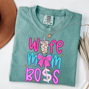 May include: A teal Comfort Colors t-shirt with the words "Wife Mom Boss" in pink and blue, with a coffee cup and bow graphic. A silver necklace with a pendant is draped across the shirt.