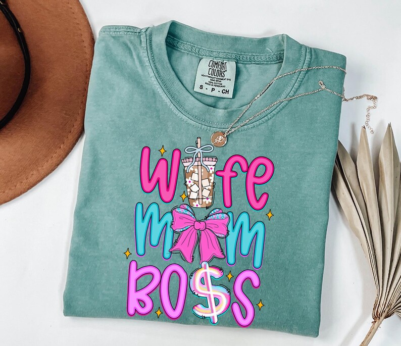 May include: A teal Comfort Colors t-shirt with the words "Wife Mom Boss" in pink and blue, with a coffee cup and bow graphic. A silver necklace with a pendant is draped across the shirt.