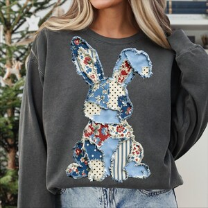May include: Dark gray sweatshirt featuring a patchwork bunny design. The bunny is made of various fabrics with floral, polka dot, and striped patterns in blue, red, and white. The sweatshirt has a crew neck and long sleeves.