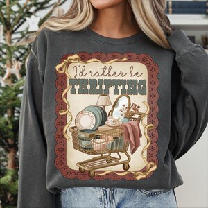 May include: Dark grey sweatshirt with a graphic that reads "I'd rather be thrifting." The graphic features a shopping cart filled with dishes, a mirror, and a lamp. The graphic is framed with a brown lace-like border and gold ribbon.