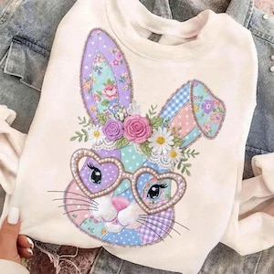 Puede incluir: Sudadera de color crema con un dise&ntilde;o de conejo de retazos. El conejo tiene gafas en forma de coraz&oacute;n, detalles florales y patrones pastel. El dise&ntilde;o incluye colores rosa, morado, azul y verde, con un estilo caprichoso y decorativo.