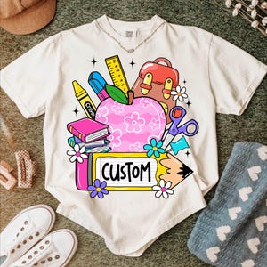 以下が含まれることがあります： クリーム色のTシャツ。カラフルな学校をテーマにしたグラフィックが特徴です。デザインには、花柄のピンク色のリンゴ、本、クレヨン、ハサミ、そして黄色のフレームに「CUSTOM」という文字が含まれています。Tシャツは他の学用品に囲まれています。