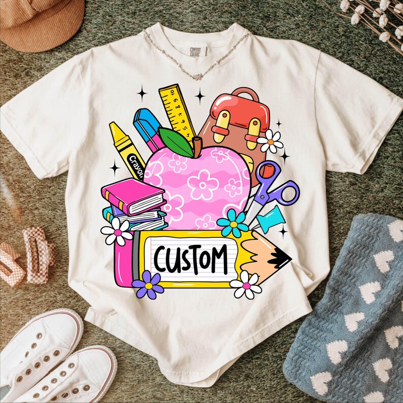 以下が含まれることがあります： クリーム色のTシャツ。カラフルな学校をテーマにしたグラフィックが特徴です。デザインには、花柄のピンク色のリンゴ、本、クレヨン、ハサミ、そして黄色のフレームに「CUSTOM」という文字が含まれています。Tシャツは他の学用品に囲まれています。
