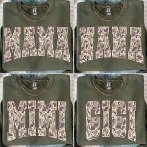 May include: Four olive green sweatshirts with gold chain necklaces. Each sweatshirt features a different name in a camouflage pattern: MAMA, NANA, MIMI, and GIGI. The sweatshirts are displayed on a denim surface.
