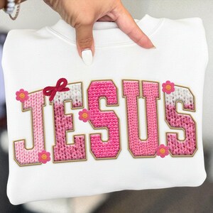 May include: White sweatshirt with the word "JESUS" in pink, knitted-style letters outlined in gold. The design includes a red bow and pink flower accents, creating a decorative and faith-based theme.