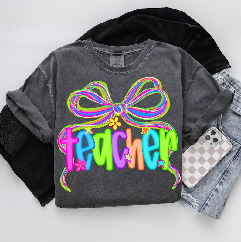 May include: Dark gray t-shirt with a colorful bow and the word "teacher" in a vibrant, multi-colored font. The design includes flowers and stars. The shirt is paired with denim shorts and a phone case.