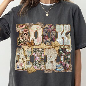 May include: Dark gray t-shirt with the words "BOOK NERD" in a collage of floral patterns, leopard print, and vintage paper. A butterfly and small books are also featured in the design.