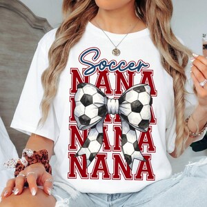 May include: White t-shirt with a soccer-themed design. The design features the word "Soccer" in blue script above the word "NANA" repeated three times in red block letters. A bow made of soccer balls is in the center.