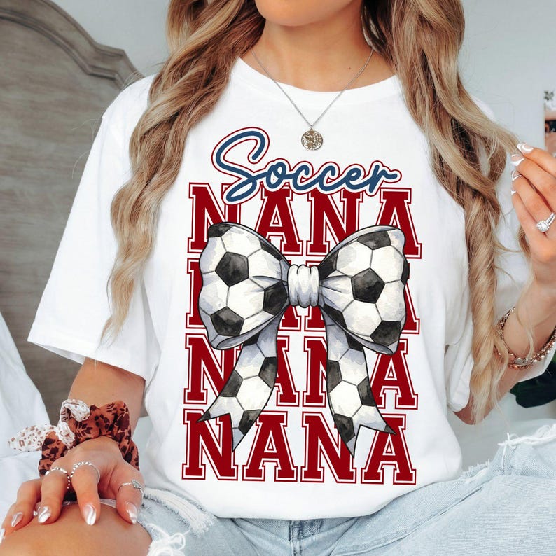 May include: White t-shirt with a soccer-themed design. The design features the word "Soccer" in blue script above the word "NANA" repeated three times in red block letters. A bow made of soccer balls is in the center.