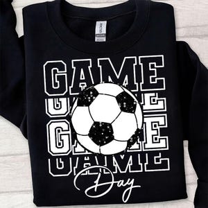 Może przedstawiać: Czarna bluza z napisem "GAME DAY" w białych literach i grafiką piłki nożnej. Piłka nożna jest czarno-biała z brokatowymi akcentami. Bluza marki Gildan.