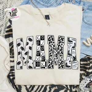 May include: A cream-coloured t-shirt featuring the word MAMA in large block letters. The letters are filled with black and white patterns including hockey pucks, goalie masks, hockey sticks, leopard print, and ice skates on a white background.