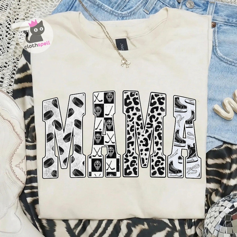 May include: A cream-coloured t-shirt featuring the word MAMA in large block letters. The letters are filled with black and white patterns including hockey pucks, goalie masks, hockey sticks, leopard print, and ice skates on a white background.