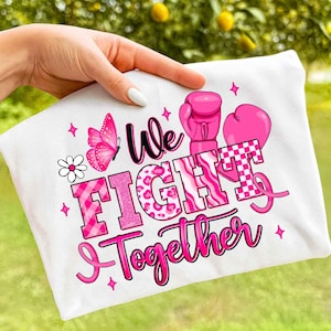 May include: White t-shirt with a pink design featuring a butterfly, boxing gloves, and the words "We Fight Together." The text is in various pink fonts and patterns, including a checkered design. Pink stars and a daisy complete the design.