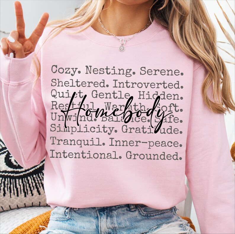 Peut inclure: Sweat-shirt rose avec le texte "Cozy. Nesting. Serene. Sheltered. Introverted. Quiet. Gentle. Hidden. Restful. Warmth. Soft. Unwind. Balance. Safe. Simplicity. Gratitude. Tranquil. Inner-peace. Intentional. Grounded." et le mot "homebody" en plus grand.