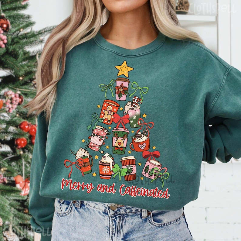 May include: Teal green sweatshirt with a Christmas tree design made of coffee cups, gingerbread men, and bows. The text "Merry and Caffeinated" is printed below the tree. A gold star tops the tree.