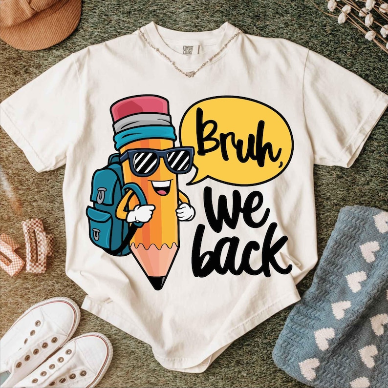 May include: Cream-colored t-shirt featuring a cartoon pencil character wearing sunglasses and a backpack. The pencil has a smiling face and the text "Bruh, we back" in a speech bubble. The shirt is on a green surface.