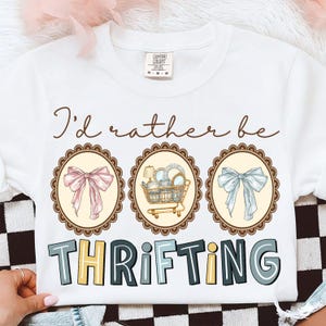 May include: White crew neck sweatshirt with the text "I'd rather be thrifting." The design includes three oval frames with illustrations of a pink bow, a shopping cart with dishes, and a blue bow. The word "THRIFTING" is in colorful block letters.
