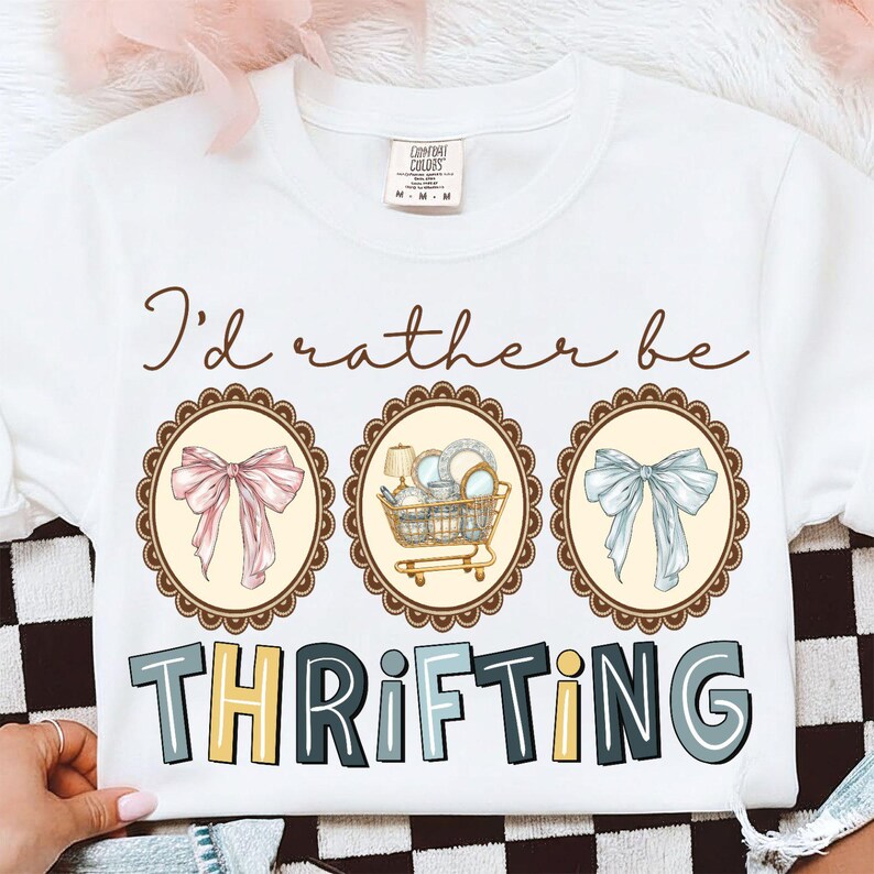 May include: White crew neck sweatshirt with the text "I'd rather be thrifting." The design includes three oval frames with illustrations of a pink bow, a shopping cart with dishes, and a blue bow. The word "THRIFTING" is in colorful block letters.