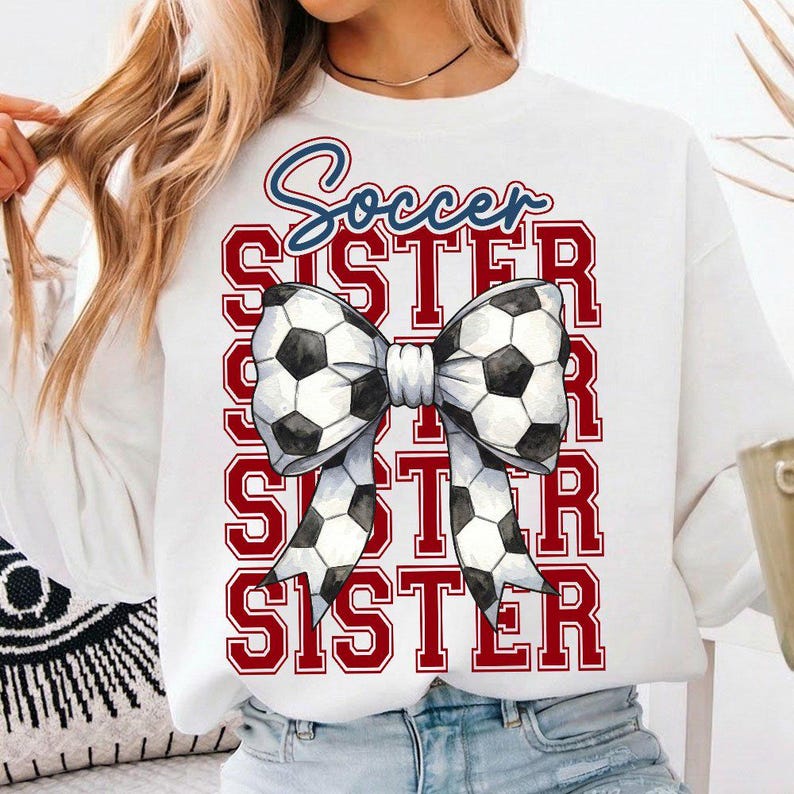 May include: White sweatshirt with the words "Soccer Sister" in blue script and red block letters. A bow made of soccer balls is centered on the front. The design is a sporty and fun graphic.