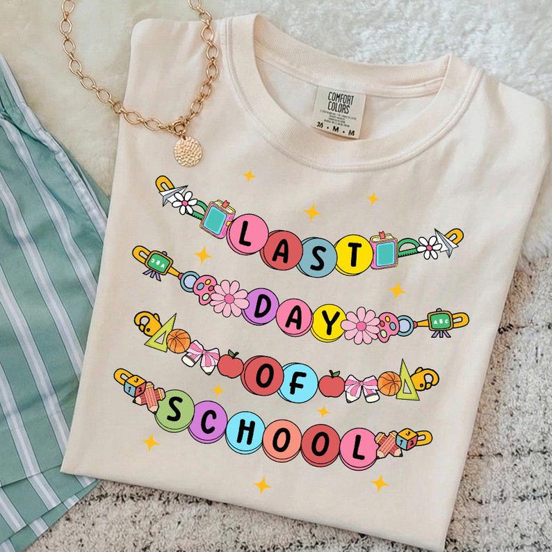 May include: Cream-colored Comfort Colors t-shirt with the text "LAST DAY OF SCHOOL" in colorful bubble letters. The design includes flowers, school supplies, and stars. A gold necklace is draped over the shirt.
