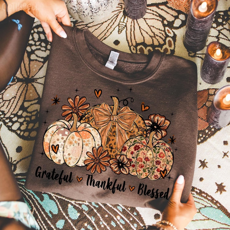 May include: A brown sweatshirt featuring a fall-themed design. The graphic includes pumpkins with floral and leopard print patterns, flowers, hearts, and the text "Grateful Thankful Blessed".