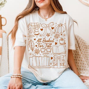 May include: Cream-colored t-shirt with a brown graphic design featuring fall-themed illustrations. The design includes a scarf, pumpkin, leaves, books, and the text "All Booked For Fall".