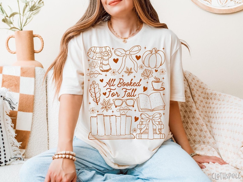 May include: Cream-colored t-shirt with a brown graphic design featuring fall-themed illustrations. The design includes a scarf, pumpkin, leaves, books, and the text "All Booked For Fall".
