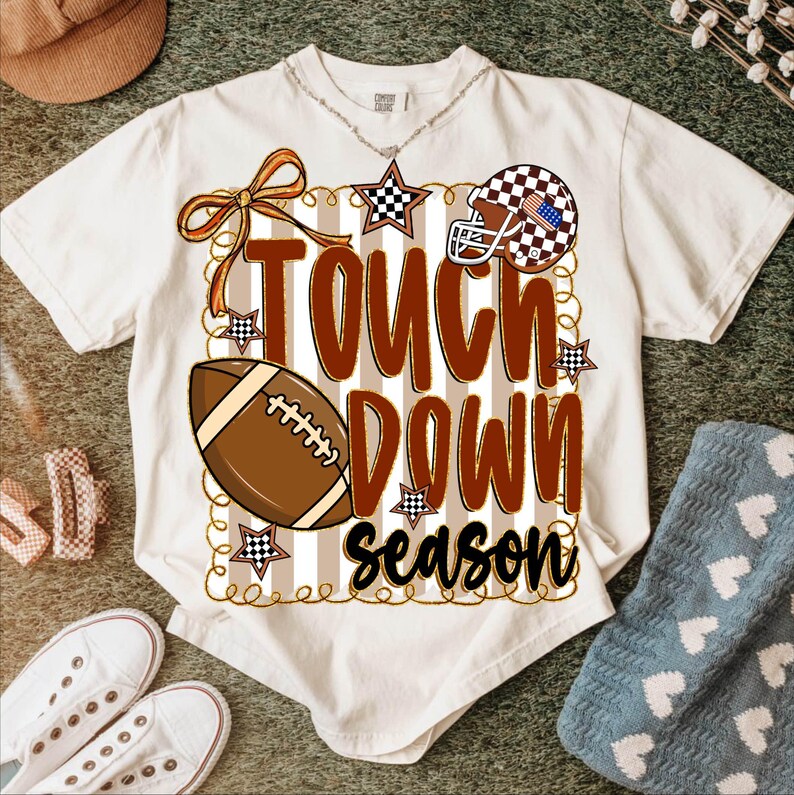 May include: Cream-colored t-shirt with a football-themed graphic. The design includes a football, a helmet with an American flag design, stars, and the words "TOUCH DOWN SEASON" in brown and black.