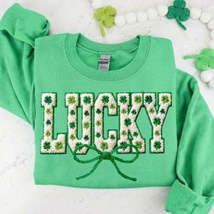 May include: Green sweatshirt with the word "LUCKY" embroidered in white and green, decorated with shamrocks. A green bow is at the bottom of the word. The sweatshirt is folded, with the sleeves visible.