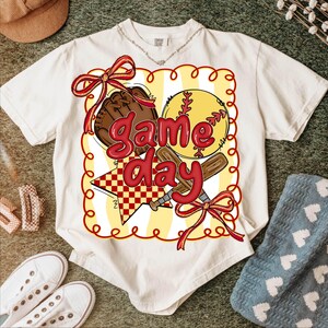 Puede incluir: Camiseta color crema con un gr&aacute;fico tem&aacute;tico de b&eacute;isbol. El dise&ntilde;o incluye una pelota de b&eacute;isbol, un guante y bates, con las palabras "Game Day" en rojo. El gr&aacute;fico est&aacute; enmarcado por un borde amarillo y rojo, con lazos rojos. La camiseta est&aacute; rodeada de accesorios informales.