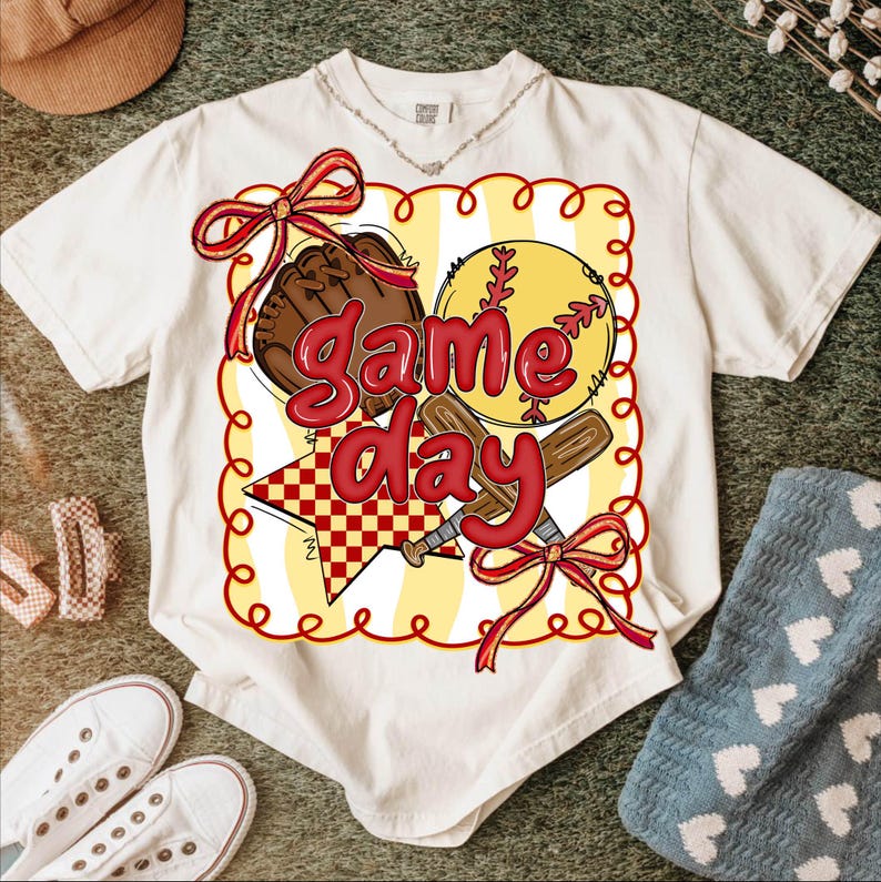 Puede incluir: Camiseta color crema con un gr&aacute;fico tem&aacute;tico de b&eacute;isbol. El dise&ntilde;o incluye una pelota de b&eacute;isbol, un guante y bates, con las palabras "Game Day" en rojo. El gr&aacute;fico est&aacute; enmarcado por un borde amarillo y rojo, con lazos rojos. La camiseta est&aacute; rodeada de accesorios informales.