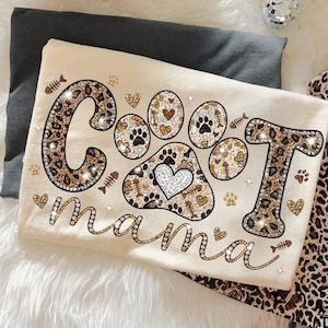 May include: Cream-colored t-shirt with the words "Cat Mama" in a decorative font. The word "Cat" is designed with leopard print and rhinestones, with a paw print design. The design includes hearts, fish bones, and paw prints.